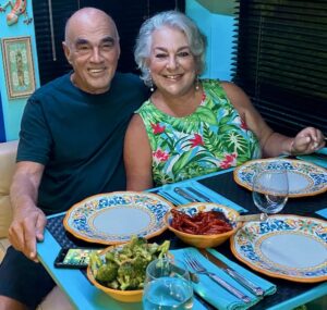 Man in black T-shirt and woman in green and pink print dress sitting at a dinette for dinner in an RV. They are the creators of Working on our Journey